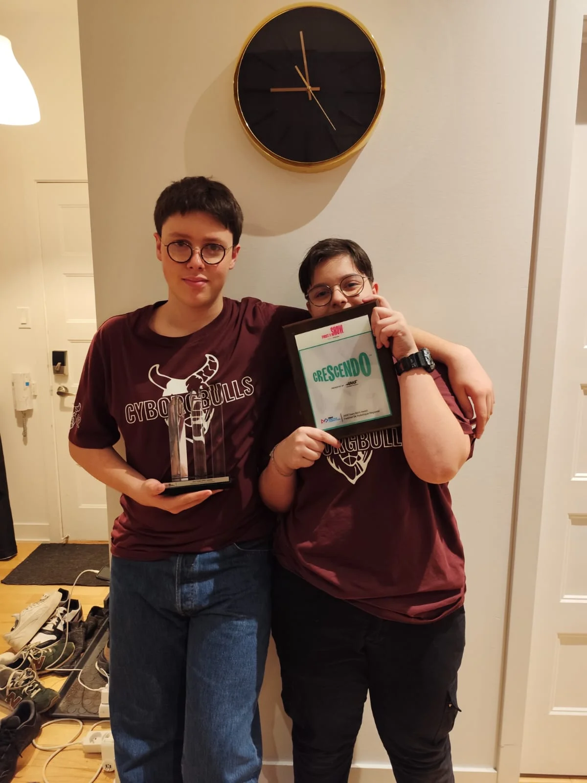 Deux membres de CyborgBulls en tee-shirts bordeaux présentent un trophée et un certificat en 2024.