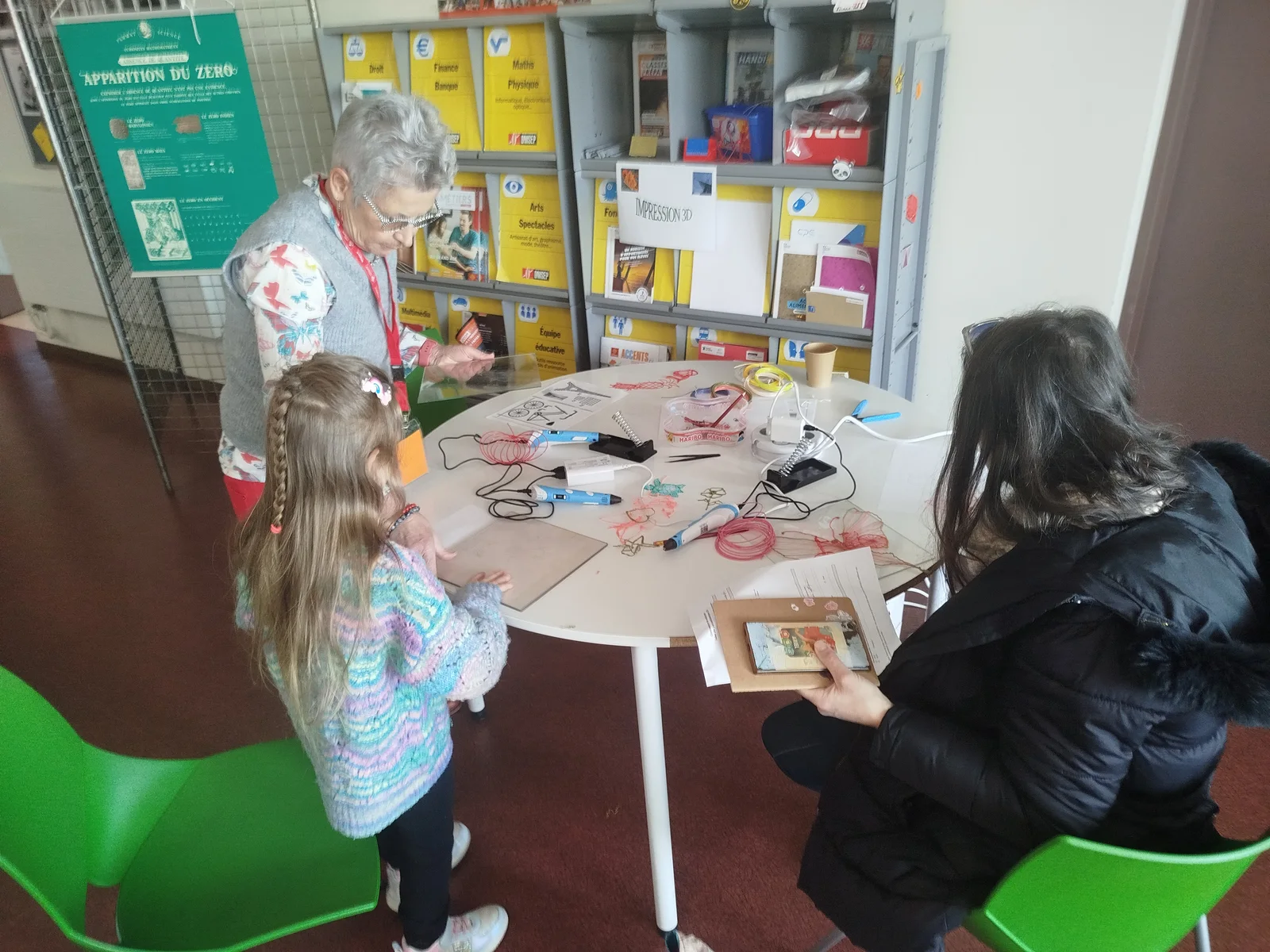 Une enfant et deux adultes autour d’une table de stylos 3D et de formes en filament.